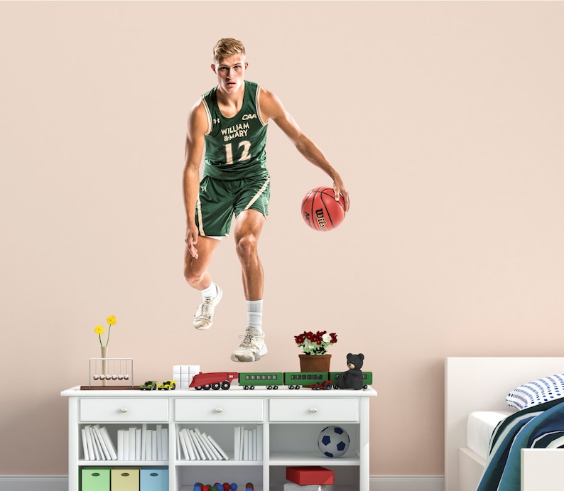 May include: A basketball player wearing a green and white jersey with the text "CAA" and "WILLIAM & MARY" and the number "12" jumps in the air while dribbling a basketball.