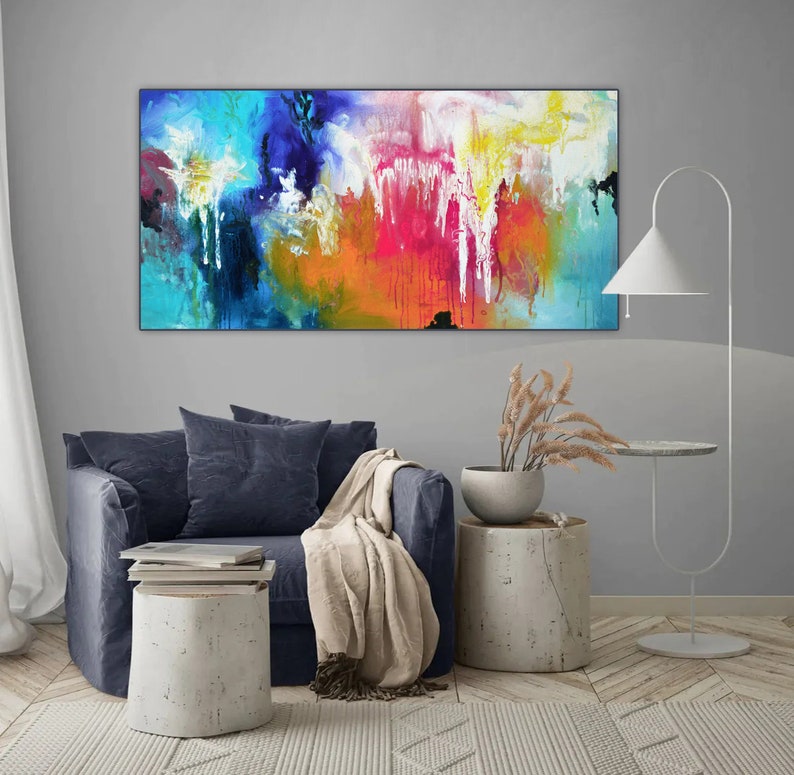 K&ouml;nnte beinhalten: Abstraktes Gem&auml;lde mit leuchtenden Farben in Blau, Gelb, Orange, Rosa und Wei&szlig;. Das Gem&auml;lde h&auml;ngt an einer grauen Wand &uuml;ber einem blauen Sofa mit einer Wolldecke.