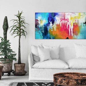K&ouml;nnte beinhalten: Abstraktes Gem&auml;lde mit leuchtenden Farben in Blau, Gr&uuml;n, Gelb, Orange, Rosa und Wei&szlig;. Das Gem&auml;lde h&auml;ngt an einer wei&szlig;en Wand &uuml;ber einem wei&szlig;en Sofa und einem runden Holz-Couchtisch.