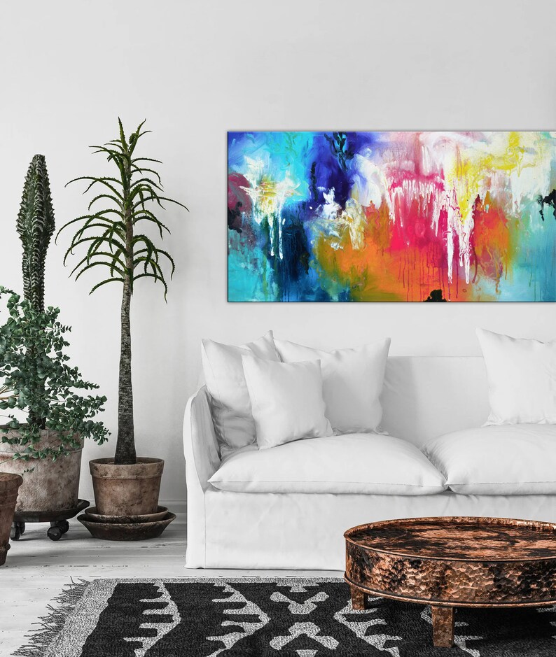 K&ouml;nnte beinhalten: Abstraktes Gem&auml;lde mit leuchtenden Farben in Blau, Gr&uuml;n, Gelb, Orange, Rosa und Wei&szlig;. Das Gem&auml;lde h&auml;ngt an einer wei&szlig;en Wand &uuml;ber einem wei&szlig;en Sofa und einem runden Holz-Couchtisch.