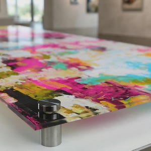 May include: A rectangular acrylic wall art panel featuring a vibrant abstract painting with bold splashes of hot pink, bright turquoise, sunny yellow, and white. The glossy panel is mounted with four circular silver metal standoffs at the corners.