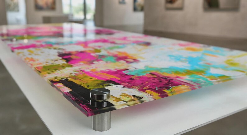 May include: A rectangular acrylic wall art panel featuring a vibrant abstract painting with bold splashes of hot pink, bright turquoise, sunny yellow, and white. The glossy panel is mounted with four circular silver metal standoffs at the corners.