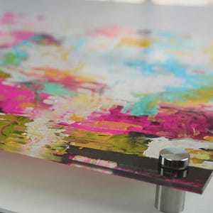 May include: A rectangular acrylic wall art panel featuring an abstract composition of vibrant pink, bright yellow, turquoise, and white paint strokes. The glossy surface is mounted with four circular silver metal standoffs at each corner.