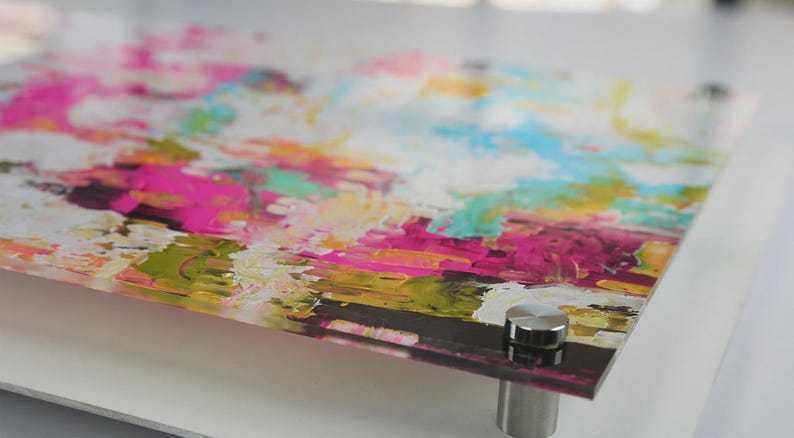 May include: A rectangular acrylic wall art panel featuring an abstract composition of vibrant pink, bright yellow, turquoise, and white paint strokes. The glossy surface is mounted with four circular silver metal standoffs at each corner.