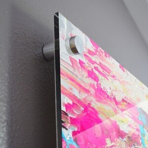 May include: A rectangular acrylic wall art piece featuring a vibrant abstract painting with thick, textured brushstrokes in shades of hot pink, white, yellow, and bright blue. The artwork is mounted to a wall using a silver metal standoff hardware mount.