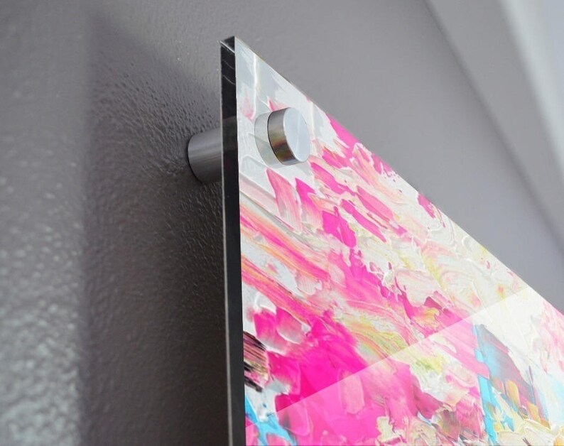 May include: A rectangular acrylic wall art piece featuring a vibrant abstract painting with thick, textured brushstrokes in shades of hot pink, white, yellow, and bright blue. The artwork is mounted to a wall using a silver metal standoff hardware mount.