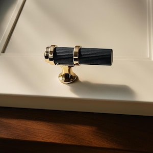 May include: A cabinet pull with a black, ribbed center section and gold-colored accents. The pull has a cylindrical shape with gold rings on each end and a gold base. The hardware is designed for use on cabinets and drawers.