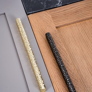 May include: Two textured cabinet pulls, one gold and one black, are displayed against a backdrop of wood and gray surfaces. The gold pull has a bright, shiny finish, while the black pull has a matte appearance. Both pulls have a linear, elongated shape.
