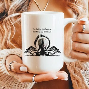 May include: White ceramic mug featuring a black illustration of a person meditating within a circle, with trees on either side. The mug has the text "The Quieter You Become, The More You Will Hear". The mug has a large handle.