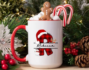 Könnte beinhalten: Eine weiße Keramiktasse mit rotem Henkel und Rand, verziert mit einem personalisierten Weihnachtsdesign. Das Design umfasst den rot-schwarz karierten Buchstaben "R" mit Weihnachtsmütze, Rentier und Schneemann. Die Tasse ist mit Marshmallows, einem Lebkuchenmann und Zuckerstangen gefüllt.