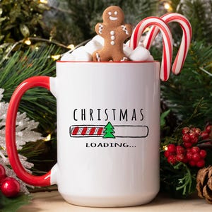 Op de afbeelding: Een witte keramische mok met een rode handgreep en rand, versierd met de woorden "CHRISTMAS LOADING..." en een kerstboomafbeelding. De mok is gevuld met marshmallows, een peperkoekmannetje en twee zuurstokken. Feestelijk vakantiethema.