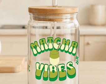 May include: A clear glass tumbler with a wooden lid and a glass straw. The tumbler features the words "Matcha Vibes" in green and white, along with a matcha drink graphic. The tumbler is designed for cold beverages.