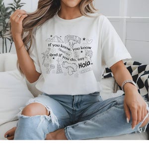 T-shirt with Venezuelan references in form of black and white doodles "el avila", "arepas", venezuelan map "guacamaya", cervical beer beer La Polar sello, "cuatro", "maracas" and Venezuelan lingo like "chama" "epale" , an oil tower, and the saying "if you know, you know And if you do, say Hola"