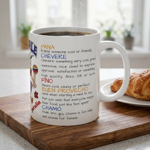 Puede incluir: Una taza de cerámica blanca con definiciones de texto coloridas de términos de jerga venezolana, incluyendo Pana, Chévere, Fino, Buen Provecho y Chamo. El diseño incluye pequeñas ilustraciones de maracas y flores junto a las definiciones.