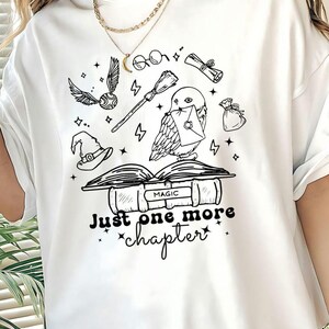 May include: White t-shirt with a black graphic of a book, owl, broom, and other magical elements. The text "Just one more chapter" is below the book. A gold necklace is worn.