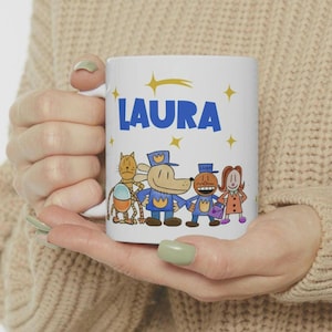 May include: White ceramic coffee mug with a blue and yellow design featuring characters from the book "The Wonderful Things You Will Be". The mug has the name "LAURA" written in blue letters on the front.