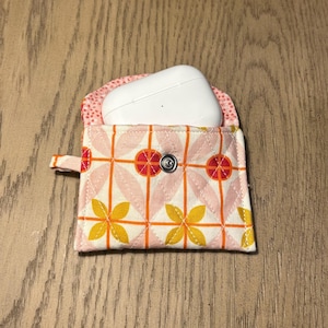 May include: A pink and yellow fabric case with a white snap closure. The case is designed with a geometric pattern and is holding a white wireless earbud case.