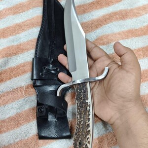 Puede incluir: Un cuchillo de caza grande con una hoja de acero pulido y un mango de material marr&oacute;n texturizado. El cuchillo tiene una guarda plateada y se combina con una funda de cuero negro.