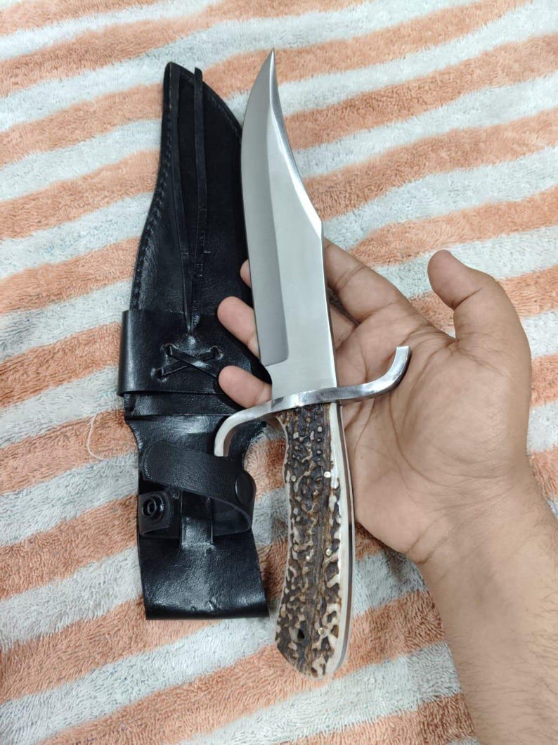 Puede incluir: Un cuchillo de caza grande con una hoja de acero pulido y un mango de material marr&oacute;n texturizado. El cuchillo tiene una guarda plateada y se combina con una funda de cuero negro.