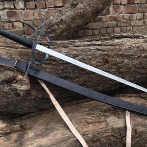 May include: A medieval-style sword with a silver blade and a black handle and guard. The sword is resting on a wooden log, with a black leather sheath. The handle has a rounded pommel.