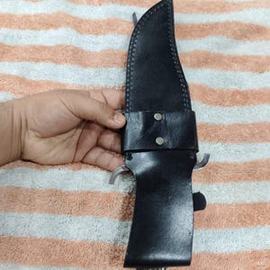 Puede incluir: Una funda de cuero negro con un cuchillo de caza grande. El cuchillo tiene una hoja plateada y un mango de hueso. La funda se asegura con una correa de cuero y dos remaches plateados.