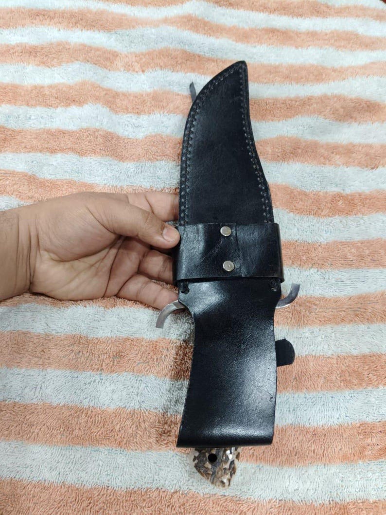 Puede incluir: Una funda de cuero negro con un cuchillo de caza grande. El cuchillo tiene una hoja plateada y un mango de hueso. La funda se asegura con una correa de cuero y dos remaches plateados.
