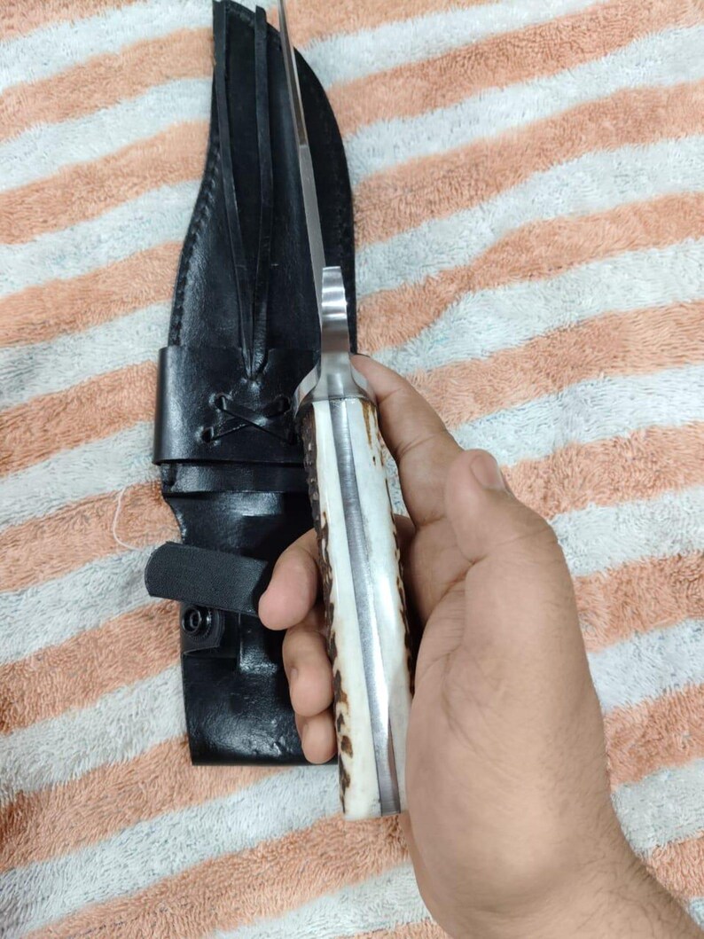Puede incluir: Un cuchillo de caza con una hoja de acero pulido y un mango de hueso y material marr&oacute;n oscuro. El cuchillo va acompa&ntilde;ado de una funda de cuero negro con trabilla para cintur&oacute;n y cierre de bot&oacute;n a presi&oacute;n.
