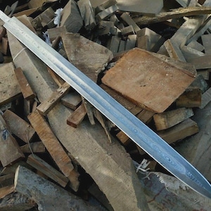 May include: A long, silver sword blade with a patterned surface rests on a pile of rough-hewn wooden blocks. The blade has a pointed tip and a flat, unsharpened tang. The wood blocks are various sizes and shades of brown.