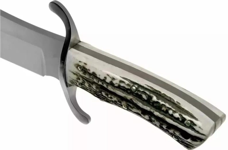 Puede incluir: Primer plano de un cuchillo de caza grande. La hoja es plateada, y el mango es una mezcla de blanco y marr&oacute;n oscuro, con una franja gris oscura en el centro. La guarda es curva y negra.