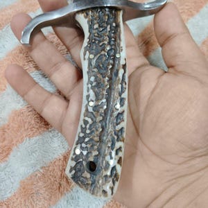 Puede incluir: Primer plano de un cuchillo de caza con una hoja de acero pulido y un mango de material texturizado marr&oacute;n claro y crema. El cuchillo tiene una guarda de color plateado.