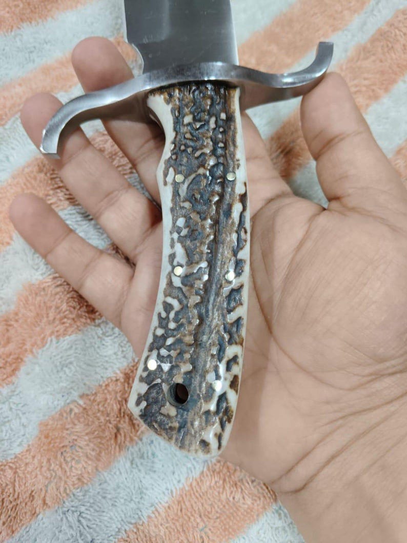 Puede incluir: Primer plano de un cuchillo de caza con una hoja de acero pulido y un mango de material texturizado marr&oacute;n claro y crema. El cuchillo tiene una guarda de color plateado.