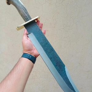 May include: A large, blue-grey,  knife with a  handle made of antler. The blade has a textured finish and a slight curve. The handle is attached to the blade with a gold-colored metal guard.
