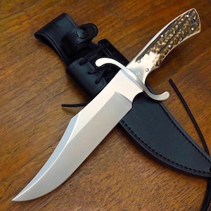 Puede incluir: Un cuchillo de caza grande de acero inoxidable con mango de hueso y funda de cuero negro. El cuchillo tiene una hoja ancha y una guarda curvada. El mango es de color marr&oacute;n claro con manchas marrones oscuras. La funda tiene un lazo para el cintur&oacute;n.