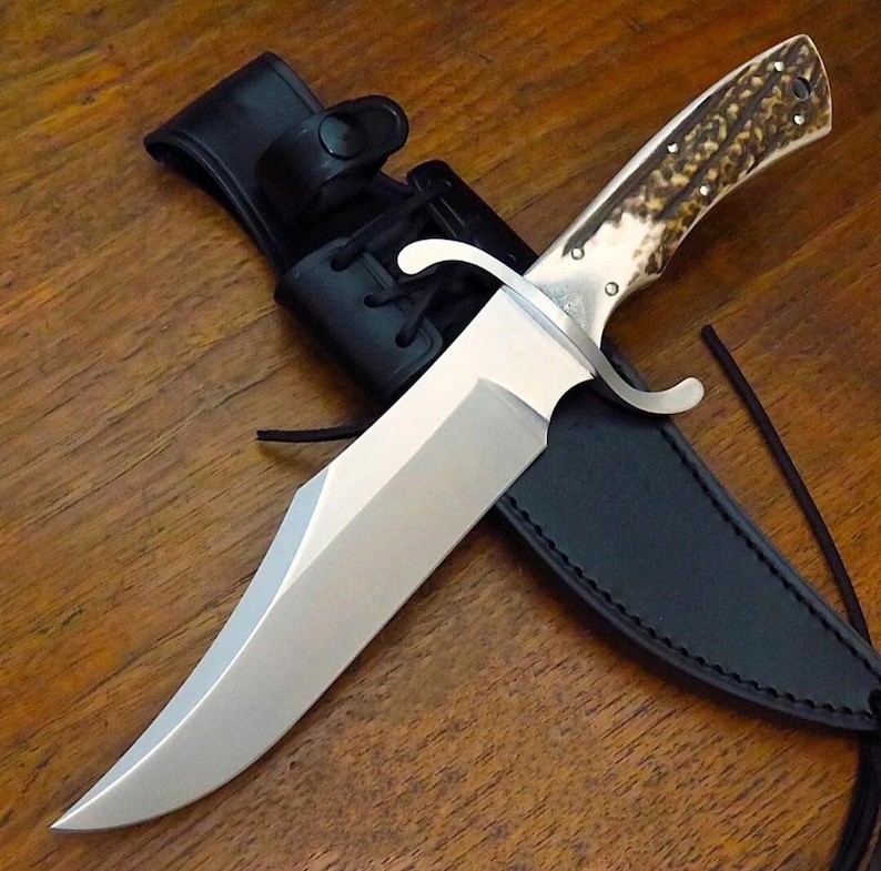 Puede incluir: Un cuchillo de caza grande de acero inoxidable con mango de hueso y funda de cuero negro. El cuchillo tiene una hoja ancha y una guarda curvada. El mango es de color marr&oacute;n claro con manchas marrones oscuras. La funda tiene un lazo para el cintur&oacute;n.