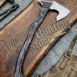 May include: A metal axe with a curved dark wood handle featuring a silver metal inlay, positioned on a wooden board with the text Vampire Hunting Store, alongside a metal hammer, pliers, a bullet, and a brown leather apron with brass buckles.