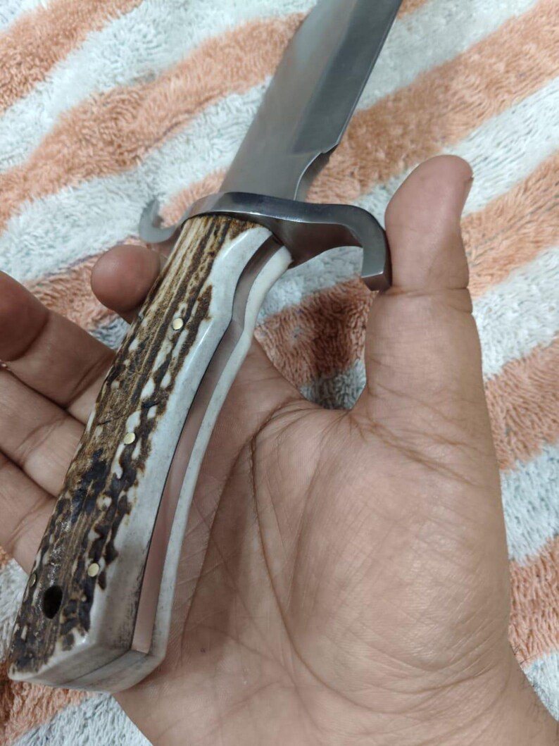 Puede incluir: Un cuchillo de caza grande con una hoja de acero inoxidable y un mango de asta y material blanco. El mango est&aacute; asegurado con remaches de lat&oacute;n. El cuchillo tiene una guarda curva.