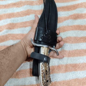 Puede incluir: Un cuchillo de caza grande con un mango de hueso y una hoja plateada curvada, asegurado en una funda de cuero negro. El mango tiene un patr&oacute;n moteado marr&oacute;n y blanco. La funda tiene una correa con cierre de bot&oacute;n.