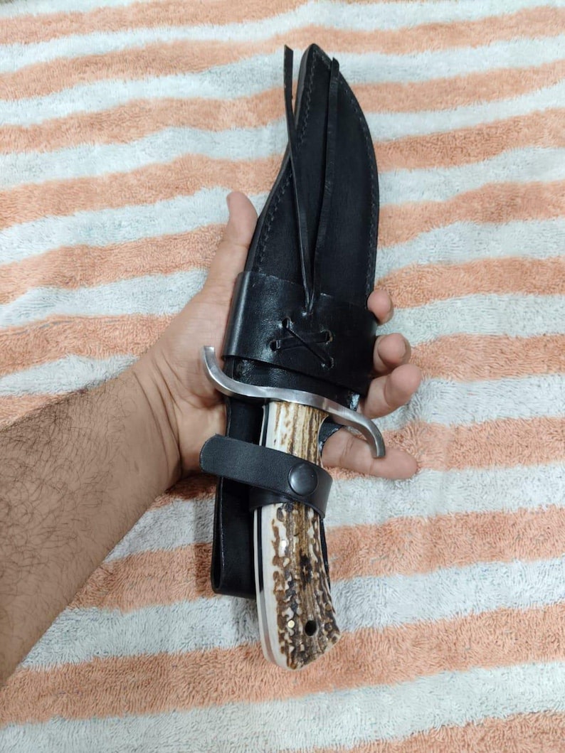 Puede incluir: Un cuchillo de caza grande con un mango de hueso y una hoja plateada curvada, asegurado en una funda de cuero negro. El mango tiene un patr&oacute;n moteado marr&oacute;n y blanco. La funda tiene una correa con cierre de bot&oacute;n.