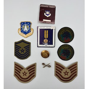 May include: A collection of military insignia, including patches, pins, and a medal. The patches feature the words "SPACE COMMAND" and "USAF CIVIL ENGINEERING." The insignia are in various colors, including blue, gold, and brown.