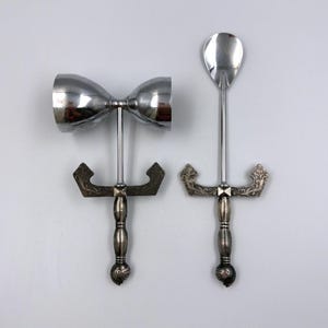 Könnte beinhalten: Ein silbernes Cocktail-Jigger und Barlöffel-Set. Der Jigger ist doppelseitig, und der Löffel hat eine tropfenförmige Schale. Beide haben verzierte Griffe mit dekorativen Elementen, ideal zum Mixen von Getränken.
