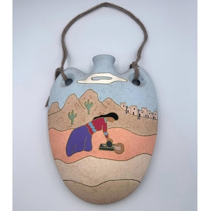 Puede incluir: Colgante de pared de cerámica ovalado con una escena desértica del suroeste. La obra de arte representa a una mujer con falda morada y top rojo, montañas, cactus y un pueblo. La pieza tiene una combinación de colores azul claro y beige, con una cuerda marrón para colgar.