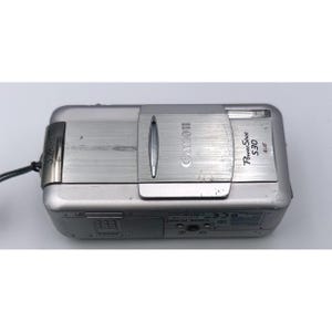 May include: Silver Canon PowerShot S30 digital camera. The camera has a retractable lens cover and a flash. The Canon logo is visible on the top. The camera has a wrist strap attached.