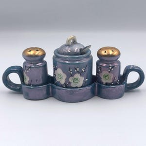 Puede incluir: Un juego de salero y pimentero de cerámica con un azucarero a juego. El juego es de color púrpura apagado con tapas doradas en los saleros y pimenteros. Diseños florales blancos decoran el juego, que incluye asas a cada lado.