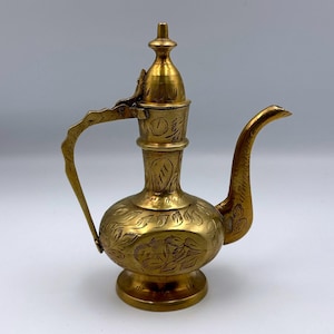 Puede incluir: Una jarra de metal dorado y ornamentado con un pico largo y un asa curvada. La jarra presenta intrincados grabados florales y de hojas en su superficie, con una pequeña tapa decorativa y una base redondeada. El diseño general sugiere una estética vintage o antigua.