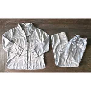 May include: A light blue satin pajama set, including a button-down shirt and drawstring pants. The shirt has a breast pocket with embroidered detailing. The set is laid out on a wooden surface.