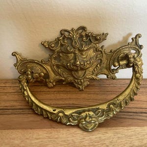 May include: A gold-toned brass drawer pull featuring an ornate, symmetrical design with a central face motif and a curved, swinging handle. The metal hardware displays intricate scrollwork and decorative detailing along the edges and the handle.
