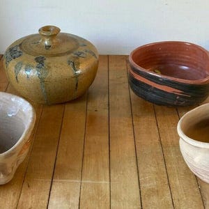 May include: Four handcrafted ceramic bowls and a lidded jar are displayed on a wooden surface. The bowls vary in shape and colour, with earthy tones of brown, tan, and red. The jar is round with a lid and a decorative knob.