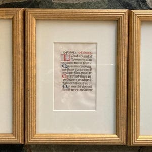 May include: Three framed pieces of art with gold-colored wooden frames. Each frame contains a rectangular piece of paper with handwritten text in black ink and red accents. The artwork is displayed on a cream-colored mat.