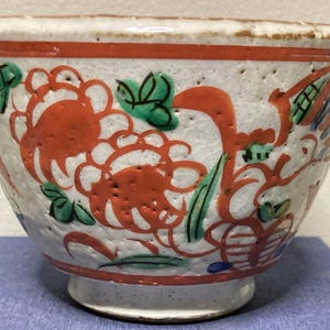 May include: A white ceramic bowl with a textured surface and a footed base. The bowl is decorated with large orange floral designs, green leaves, and accents of blue. A red band encircles the top and base. This decorative bowl is a unique piece.