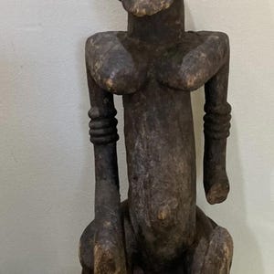 Puede incluir: Escultura de madera marrón oscuro de una figura sentada con cabeza redondeada, senos prominentes y brazos doblados a la altura de los codos. La figura está sentada sobre una base de madera, mostrando un aspecto envejecido.